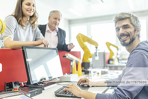 Zufriedene Kollegen beobachten lächelnden Mann bei der Arbeit am PC in der Roboterfabrik