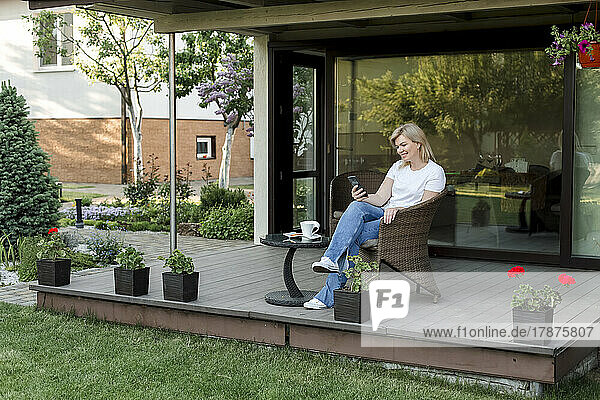 Smiling woman using smart phone sitting on chair at porch