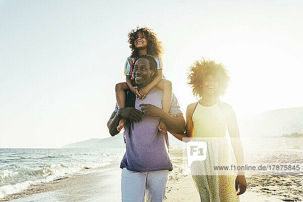 Happy man carrying girl on shoulder walking with daughter at beach on sunny day