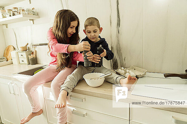 Mädchen und Bruder halten Shaker über Schüssel und sitzen zu Hause auf der Küchentheke