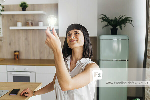 Smiling woman holding illuminated light bulb in kitchen at home