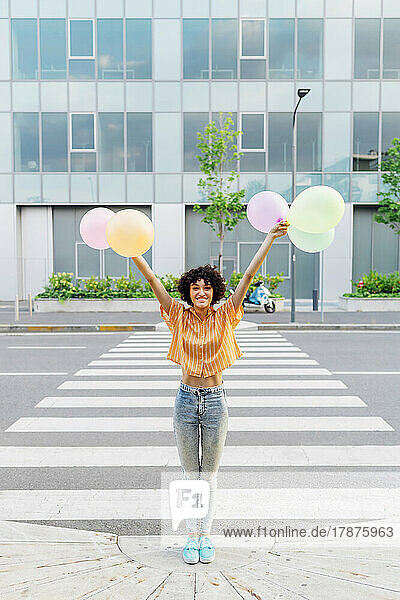 Smiling woman with arms raised holding balloons at zebra crossing on street
