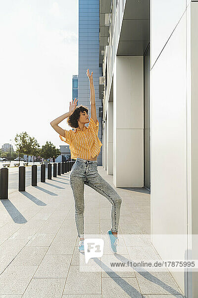 Carefree woman with hand raised dancing on sidewalk by building