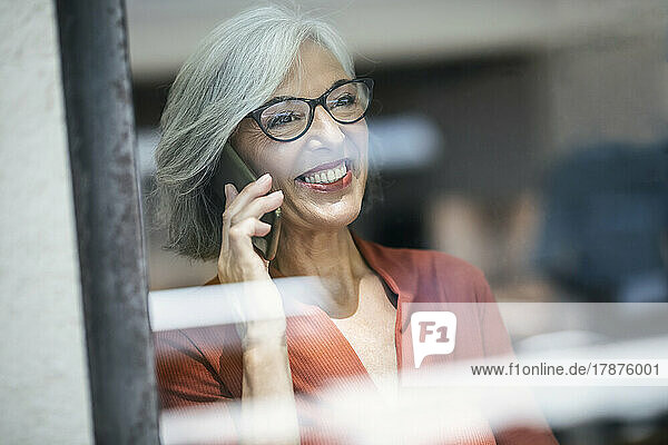Smiling senior businesswoman talking on phone seen through glass window