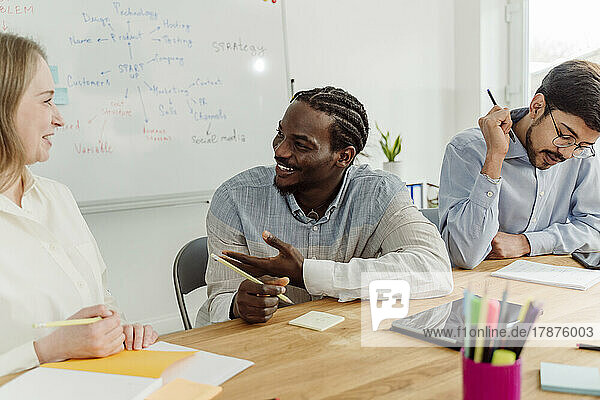 Smiling businesswoman discussing with businessman sitting by colleague in office