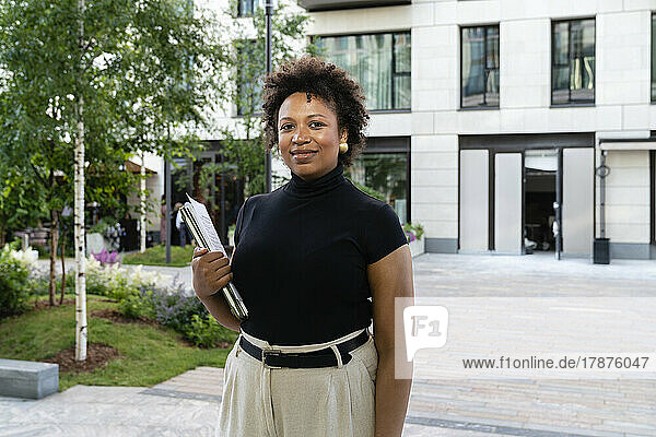 Smiling businesswoman with laptop standing in front of office building