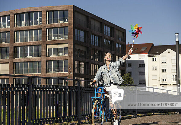 Happy woman with pinwheel toy riding bicycle by railing