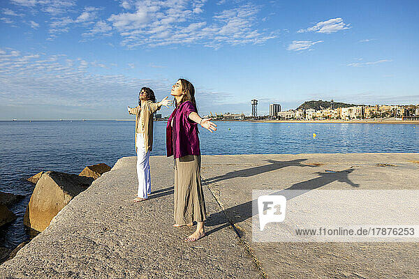 Young couple with arms outstretched enjoying sunshine on pier