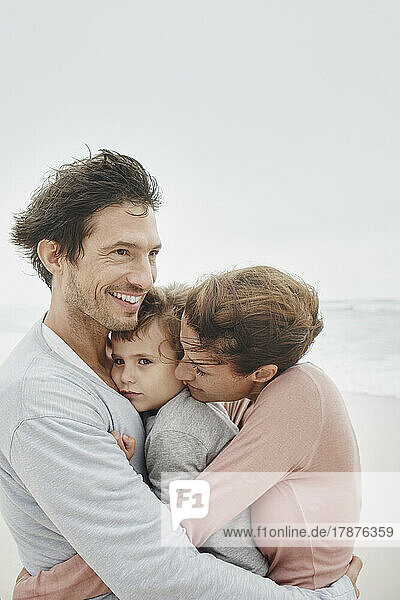 Fürsorgliche Eltern umarmen und küssen ihren Sohn am windigen Strand