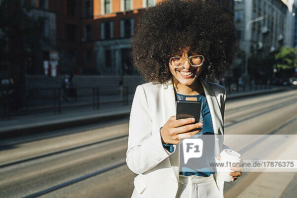 Smiling businesswoman with smart phone and coffee cup crossing street on sunny day