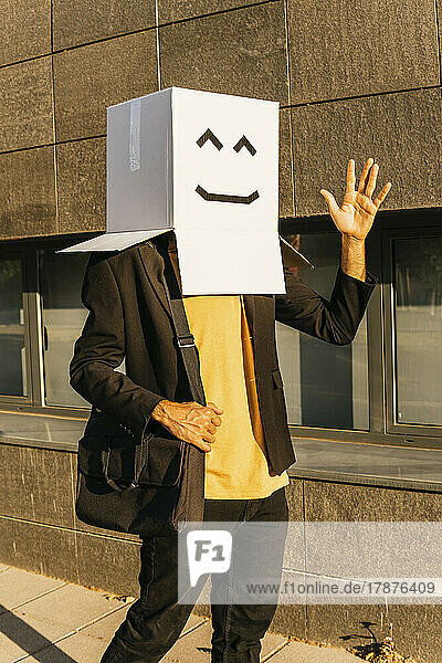 Businessman wearing box with smiley face waving hand on footpath