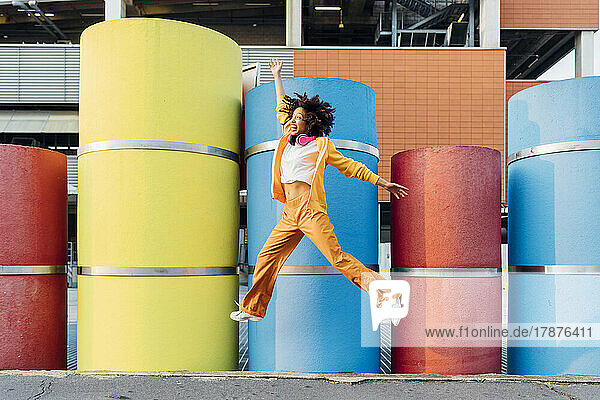 Happy young woman jumping with hand raised in front of colorful pipes