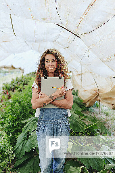 Smiling farm worker with clipboard standing in greenhouse