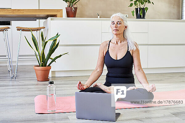 Reife Frau sitzt zu Hause im Lotussitz auf der Matte am Laptop