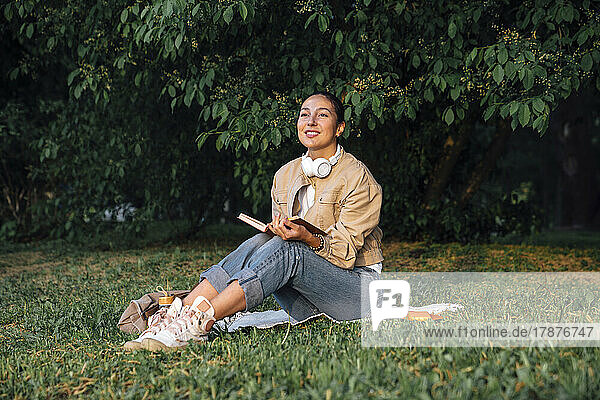 Lächelnde Frau sitzt mit Buch und kabellosen Kopfhörern im Park