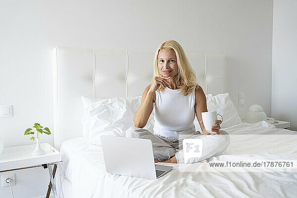 Woman holding tea sitting with hand on chin by laptop on bed