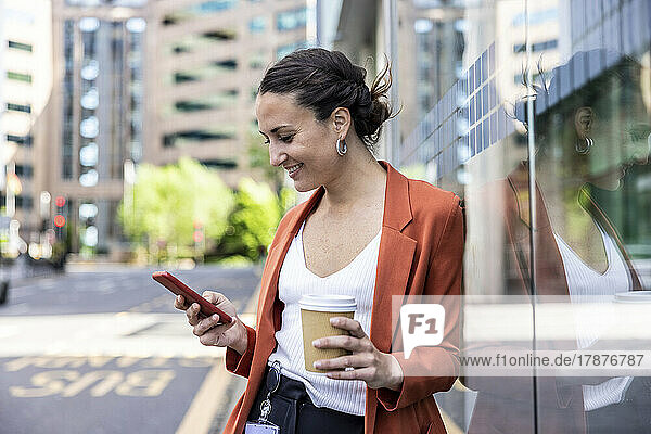 Smiling businesswoman holding disposable cup using smart phone leaning on glass wall