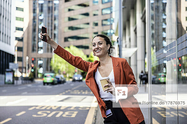 Happy businesswoman holding disposable cup and mobile phone hailing ride in city