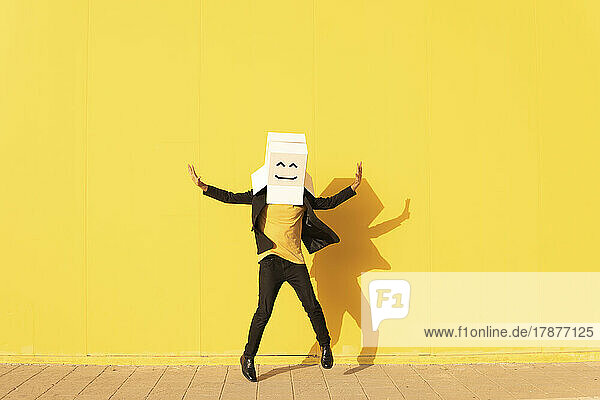 Happy man wearing box with smiley face jumping in front of yellow wall