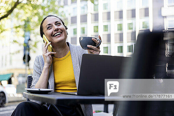 Happy businesswoman holding coffee cup talking on mobile phone at sidewalk cafe