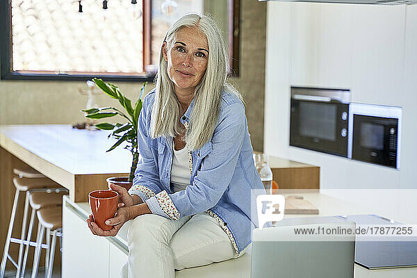 Reife Frau mit Kaffeetasse sitzt auf der Küchentheke