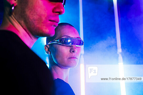 Young woman wearing futuristic glasses by man near illuminated background