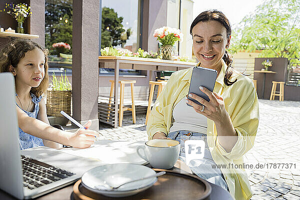 Smiling woman using smart phone sitting by daughter at sidewalk cafe