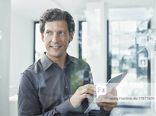 Smiling businessman with tablet PC seen through glass in office