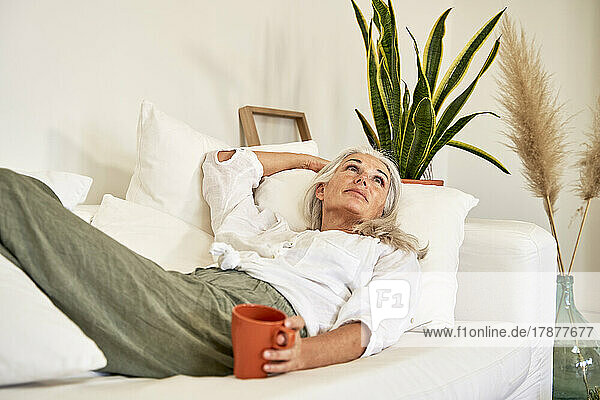 Thoughtful woman with mug lying on sofa