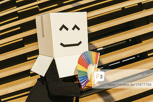Businessman wearing box with smiley face holding rainbow color hand fan on sunny day