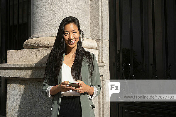 Smiling woman with smart phone standing in front of building
