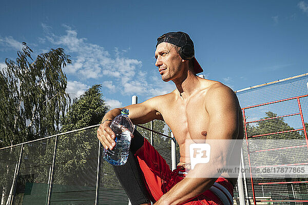 Sportsperson listening music through wireless headphones sitting with water bottle at park on sunny day