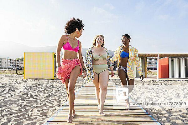 Happy friends in swimwear walking together on boardwalk at beach