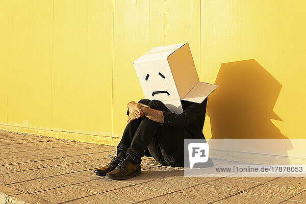 Man wearing box with sad face sitting on footpath