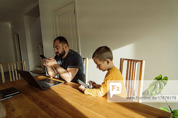 Man using smart phone sitting by son with tablet PC at home