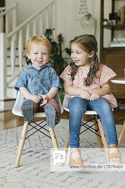 Smiling brother (12-17 months) and sister (2-3) sitting on chairs at home
