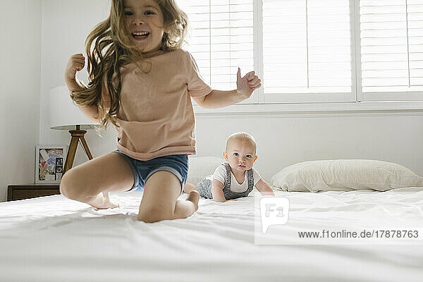 Girl (2-3) jumping on bed with baby brother (6-11 months) in background