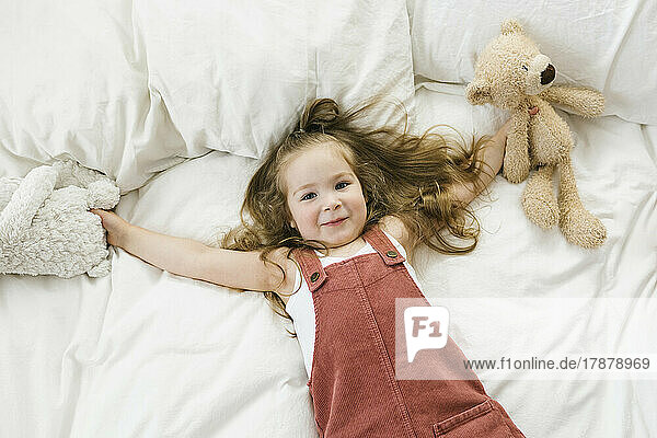 Overhead view of smiling girl (2-3) lying on bed with stuffed toys