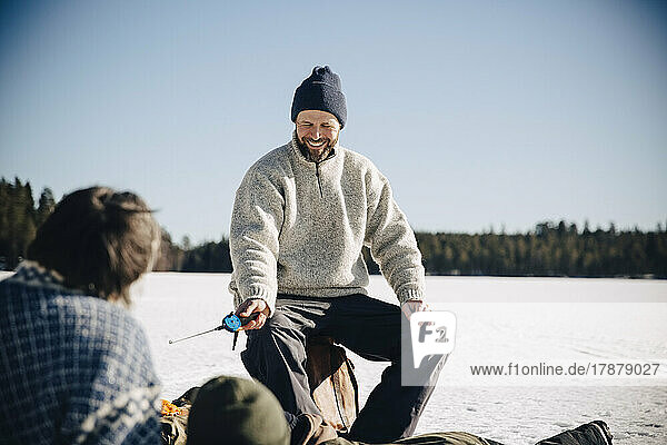 Happy mature man holding fishing rod sitting with friends during sunny day in winter