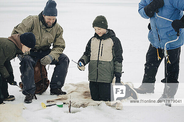Happy boy kneeling by men and friend while fishing at frozen lake during winter