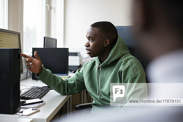 Male trainee in hooded shirt pointing at desktop screen while discussing in office