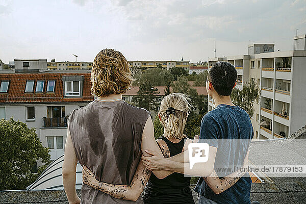 Rear view of male and female friends standing with arms around on rooftop