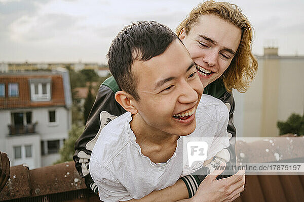 Cheerful young man embracing male friend while enjoying together on rooftop