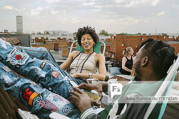 Happy young woman looking at friend sitting on chair at rooftop