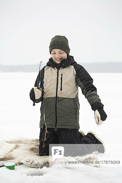 Glücklicher Junge in warmer Kleidung kniend beim Angeln auf einem zugefrorenen See