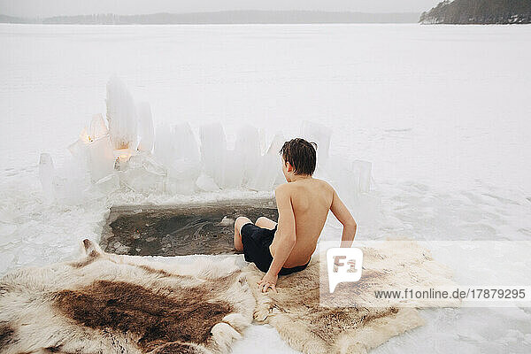 Rear view of shirtless boy sitting with legs in cold water at frozen lake