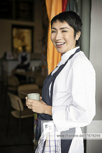Side view of happy business owner holding cup while standing at entrance of restaurant