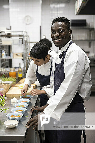 Porträt eines lächelnden Kochs  der neben einer Kollegin steht  die in einer Großküche Lebensmittel garniert