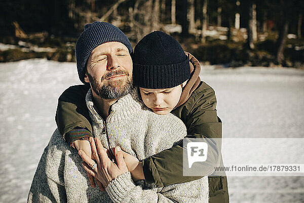 Boy in warm clothing hugging father with eyes closed during sunny day in winter