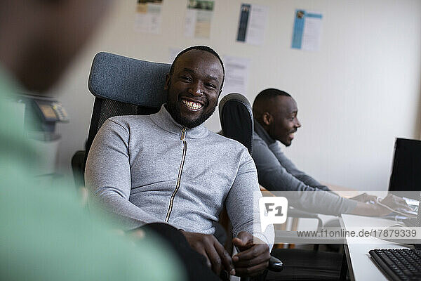 Porträt eines fröhlichen Geschäftsmannes auf einem Stuhl sitzend mit Mitarbeitern im Büro